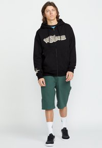Sweat à zip noir avec un graphisme "Volcom", shorts cargo verts, chaussettes blanches et baskets noires, se tenant devant un fond clair.