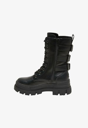 Persoon die zwarte leren broek en laarzen draagt, een laars aanpast terwijl hij uit een auto stapt op een stoep in zonlicht.