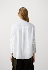 Chemise blanche texturée à manches longues avec un pli au dos, ourlet arrondi, et poignets boutonnés, associée à un pantalon foncé.
