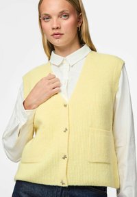 Mlada žena s dugom plavom kosom nosi svijetložutu prsluk s gumbima preko bijele košulje s ovratnikom i tamnim trapericama, na bijeloj pozadini.