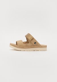 Lys brun semsket sandle med justerbar stropp, åpen tå og teksturerte detaljer. Har en lett kremfarget gummisåle med et mønster for godt grep.