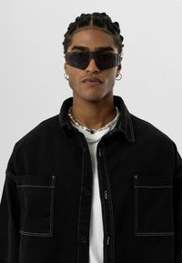 Zwarte denimjas met contrasterende witte stiksels en een borstzak, gedragen over een wit shirt. De persoon draagt donkere zonnebrillen en gelaagde kettingen.