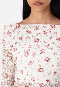 Mujer que lleva un vestido blanco de encaje con escote off-shoulder, adornado con bordados de flores en tonos rosa y rojo, visible desde el cuello hasta la mitad del torso.