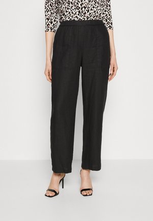 Kvinde iført sorte, højtaljede bukser med vide ben og forlommer foran samt sorte, åbne højhælede sandaler.