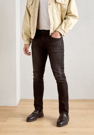 Homme portant un jean slim noir, une veste beige, une chemise blanche et des chaussures en cuir noires, debout sur un sol en bois contre un mur uni.