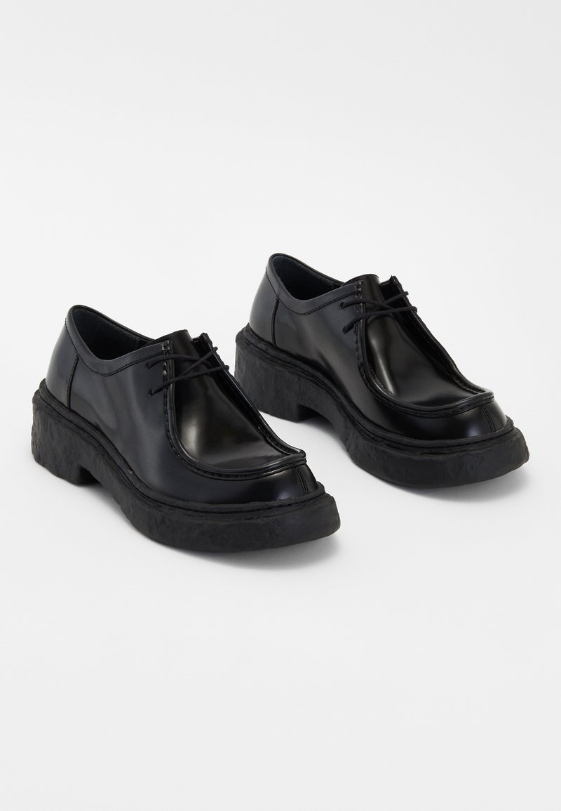 Zapatos de cuero negro con punta redonda, diseño de cordones y suela de goma negra texturizada, con costuras en contraste a lo largo de los bordes.
