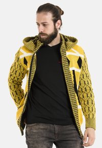 Gelber Strickcardigan mit schwarz-weißen Mustern, ausgestattet mit einer strukturierten Kapuze und einem Reißverschluss. Rippbündchen und Saum.