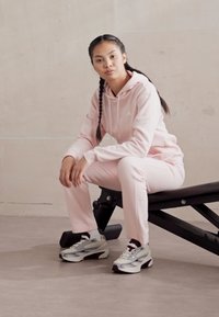 Moletom rosa claro e calças de entrecot correspondentes feitas de um tecido suave, sentados num banco preto, combinados com ténis atléticos cinza e branco.