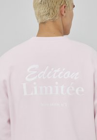 Sweat-shirt rose clair en tissu doux, avec du texte en relief blanc indiquant "Édition Limitée hors série n°1" dans le dos.