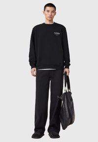 Man wearing black sweatshirt with white logo, black wide-leg pants, black shoes, holding large black tote bag against plain background.