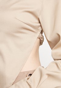 Haut beige à manches longues avec découpes élastiques sur les côtés laissant apparaître la peau, associé à un pantalon beige taille haute avec un bouton visible.