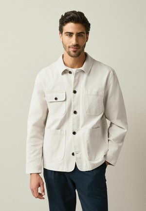 Man with short dark hair wearing a light beige button-up jacket with four pockets and dark trousers standing against plain background.