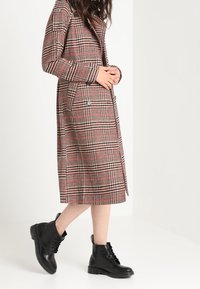 Cappotto a quadri, lungo fino al ginocchio, rosso, nero e bianco, con tasche laterali e dettagli con bottoni, abbinato a stivali alla caviglia in pelle nera con lacci.