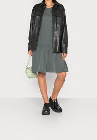 Camisa chaqueta de cuero negro sobre un vestido verde con pliegues, combinada con zapatos slip-on negros de plataforma y un bolso ligero verde.