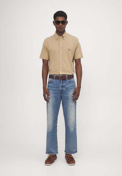 Camisa polo de manga curta beje, jeans azuis desgastados, cinto castanho e sapatos de barco castanhos, com óculos de sol escuros, posando contra um fundo branco.