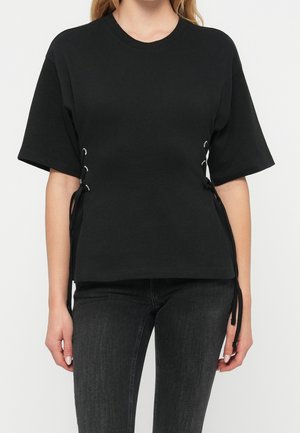 Femme portant un haut noir à manches courtes avec des détails à lacets sur les côtés, associé à un jean gris foncé sur un fond blanc uni.