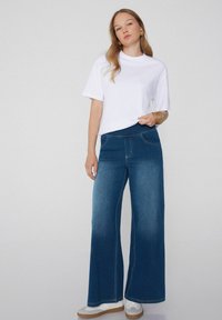 Pantalones de denim de pierna ancha con un lavado azul medio, bolsillos frontales y costuras en contraste, combinados con una camiseta blanca de manga corta.
