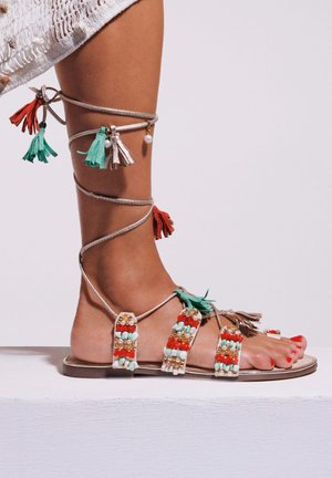 Sandales plates en perles avec une base beige, des pompons colorés en rouge, vert et blanc, présentant des embellissements complexes autour des lanières.
