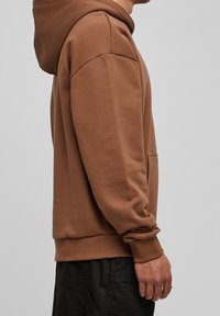 Brun hoodie med kängurufickor, huva och långa ärmar. Slät textur med ribbade mudd och nederkant. Sidoprofilvy.