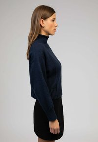 Pull en maille marine avec un col montant et un ourlet côtelé, associé à une mini-jupe noire texturée. La vue de côté met en avant le design et l'ajustement.