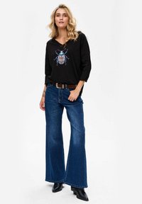 Woman with blonde hair wearing a black long-sleeve shirt featuring a blue beetle graphic, blue wide-leg jeans, and black pointed boots.