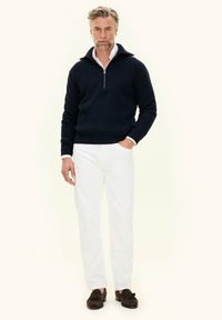Uomo che indossa un maglione navy a mezza zip, una camicia bianca con colletto, pantaloni bianchi e mocassini con nappine marroni, in piedi con una mano in tasca.