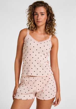 Femme aux cheveux bouclés portant un caraco rose clair à pois et un short assorti sur fond blanc uni.