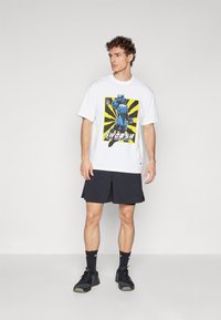 White t-shirt featuring a colorful graphic print of a superhero and the text "SWOOSH." Worn with black athletic shorts and black shoes.