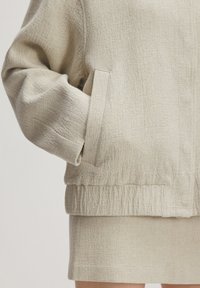 Texturierte beige Jacke mit seitlichen Taschen und geripptem Saum, kombiniert mit einem passenden Rock. Beide Kleidungsstücke weisen ein dezentes, gewebtes Muster auf.