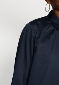 Námořnická košile s hladkým povrchem a jemným leskem. Má volný střih, mírně nabírané rukávy a klasický výstřih.