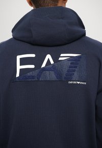 Tamsiai mėlynas hoodie, kuriame nugarinėje pusėje didelis "EA7" logotipas su tekstūruotu mėlynos spalvos akcentu ir "Emporio Armani" etikete apatinėje dešinėje.
