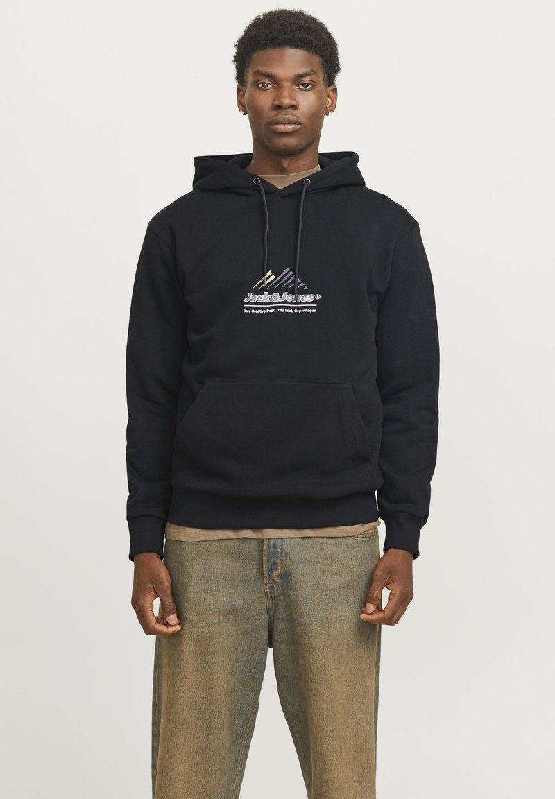 Jeune homme portant un sweat à capuche noir Jack & Jones et un jean délavé vert brun, debout contre un fond clair uni.