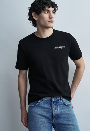 Νεαρός άνδρας με σγουρά μαλλιά που φορεί μαύρο μπλουζάκι Off-White και μπλε τζιν, στέκεται μπροστά σε ένα απλό ανοιχτό γκρι φόντο.