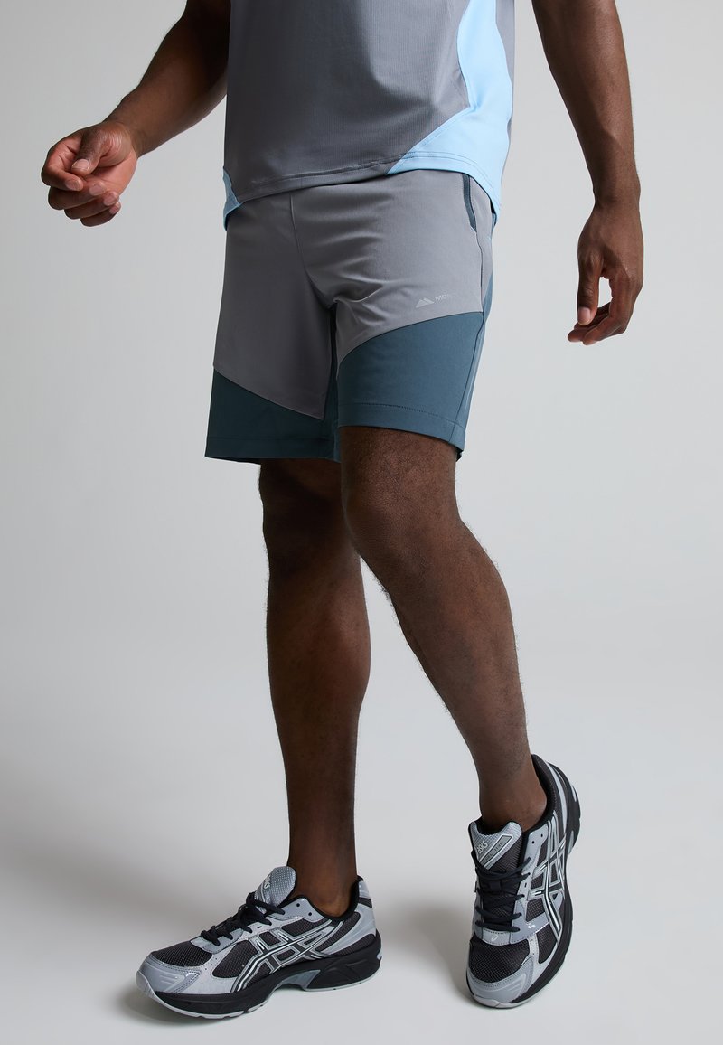 Homme portant un short de sport gris et bleu sarcelle, des chaussures de course grises et noires, debout avec les genoux légèrement pliés sur un fond clair.