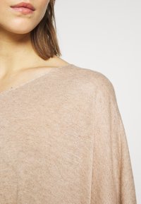 Beżowy sweter z dzianiny z szerokim dekoltem w kształcie litery V i miękkim, teksturowanym materiałem. Krój jest luźny i delikatnie opada na ramionach.