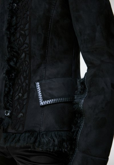 Veste en daim noire avec rabat de poche en cuir texturé, panneau décoratif en dentelle et bordure en fourrure le long des bords et des poignets.