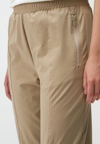 Beige, lichte broek met een elastische tailleband, met een ritszak aan de zijkant, een gladde textuur en subtiele stikdetails.