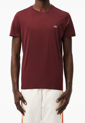 Homme portant un t-shirt bordeaux uni à manches courtes avec un petit logo crocodile vert sur la poitrine et un pantalon blanc avec des rayures orange.