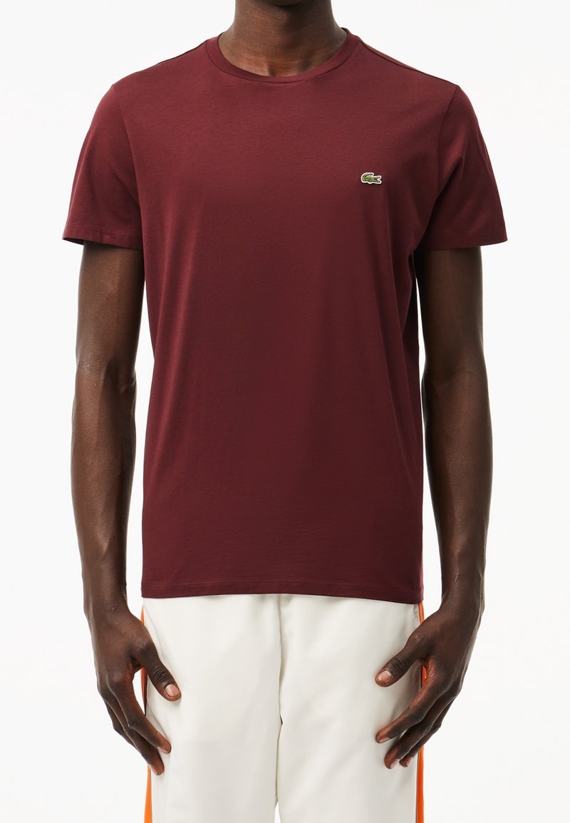 Homme portant un t-shirt bordeaux uni à manches courtes avec un petit logo crocodile vert sur la poitrine et un pantalon blanc avec des rayures orange.