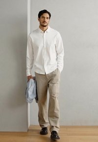 Witte button-down shirt, beige broek, houdt een lichtblauwe jas vast, zwarte schoenen. Casual, getailleerde snit; gladde stof met een schoon ontwerp.