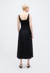 Άτομο με κοντά ξανθά μαλλιά που φοράει μαύρο μίντι φόρεμα χωρίς μανίκια και μαύρα σανδάλια με τακούνι, στέκεται μπροστά σε απλό φωτεινό φόντο.