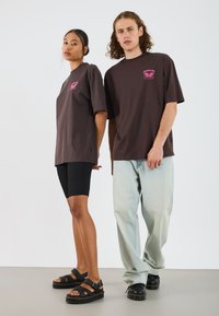 Brown oversized t-shirts with pink graphic, black bike shorts, and chunky black sandals. Models stand against a white background.
