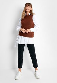 Bruine ribgebreide vest over een wit overhemd, gecombineerd met zwarte tapered pants en witte sneakers. Eenvoudig en gelaagd outfitontwerp.