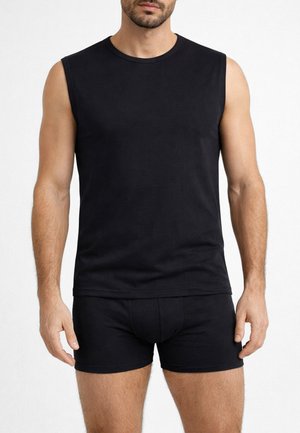 Uomo che indossa una canotta nera senza maniche e boxer neri in piedi davanti a uno sfondo bianco semplice.