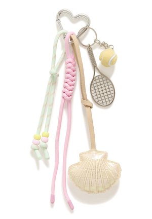 Femme blonde portant un haut blanc sans manches et un pantalon beige, avec un grand sac à bandoulière en crochet marron orné d'un porte-clés coquillage.