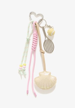 Femme blonde portant un haut blanc sans manches et un pantalon beige, avec un grand sac à bandoulière en crochet marron orné d'un porte-clés coquillage.