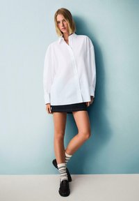 Camicia bianca oversize con bottoni e maniche lunghe, abbinata a una gonna mini in denim scuro, calze a righe e scarpe nere, su uno sfondo blu.