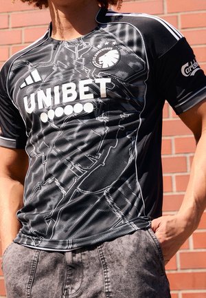 Personne portant un maillot de football noir du FC København avec les logos blancs Adidas et UNIBET, debout contre un mur de briques, les mains dans les poches.