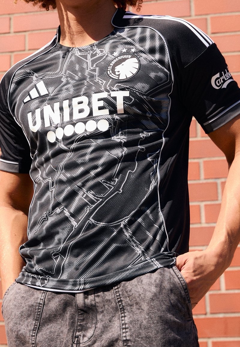 Personne portant un maillot de football noir du FC København avec les logos blancs Adidas et UNIBET, debout contre un mur de briques, les mains dans les poches.