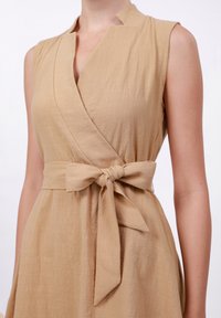 Beige linen dress with a wrap design, sleeveless cut, and a tie belt at the waist. Textured fabric with a subtle sheen.